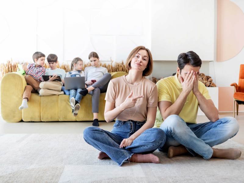 Frau und Mann sitzen auf dem Boden, im Hintergrund sieht man vier Kinder auf einem Sofa sitzen.