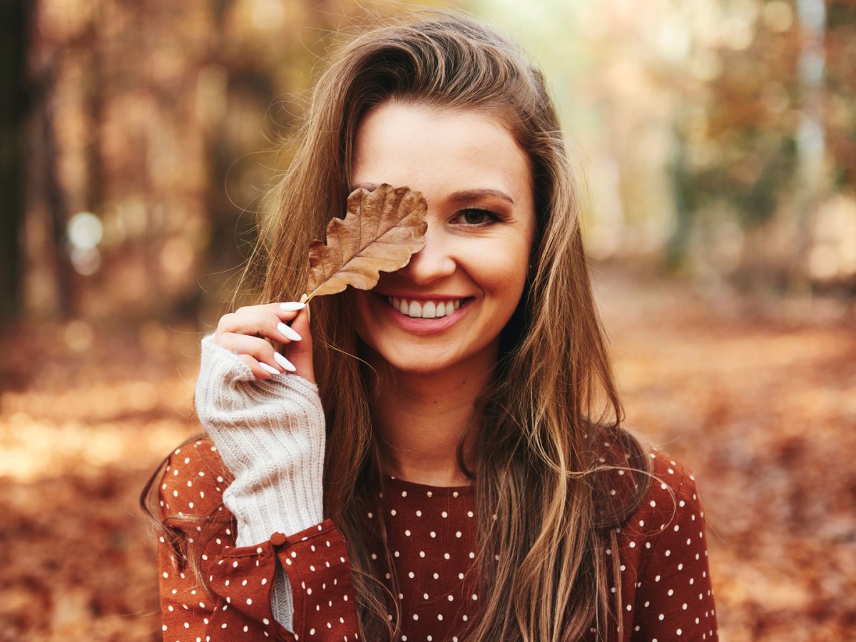 Br&uuml;nette Frau, die dem Herbsttyp entspricht, steht in herbstlichem Wald und h&auml;lt ein Laubblatt vor ihr Auge