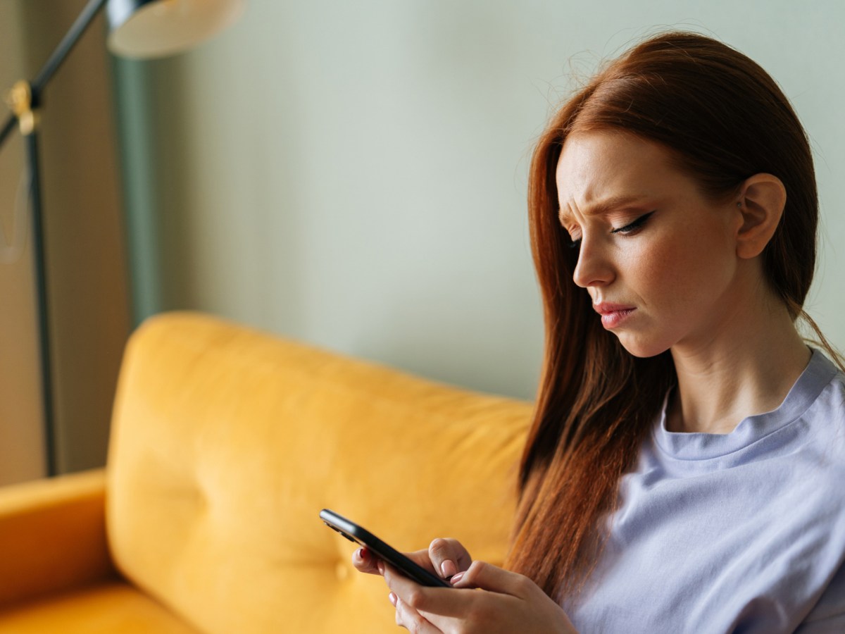 Rothaarige Frau auf gelber Couch, die ein Handy in der Hand hält und skeptisch auf den Bildschirm schaut