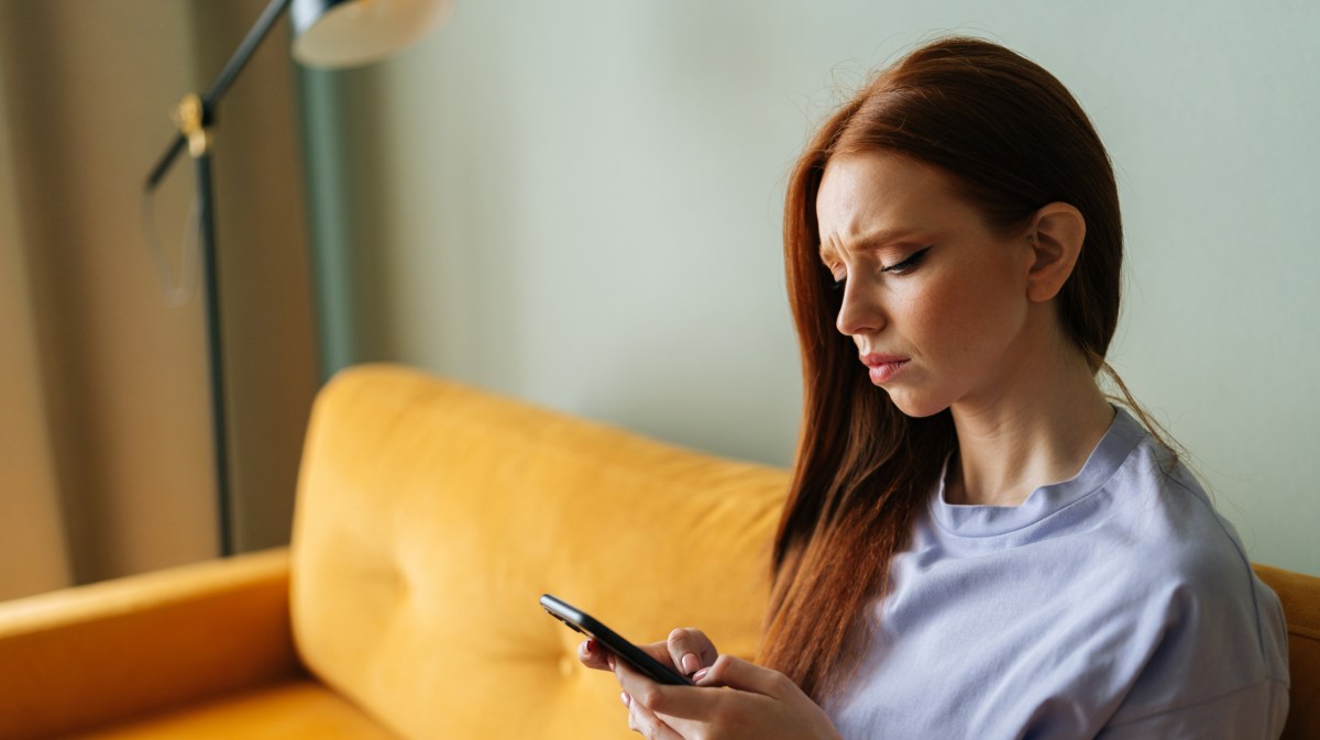 Rothaarige Frau auf gelber Couch, die ein Handy in der Hand hält und skeptisch auf den Bildschirm schaut