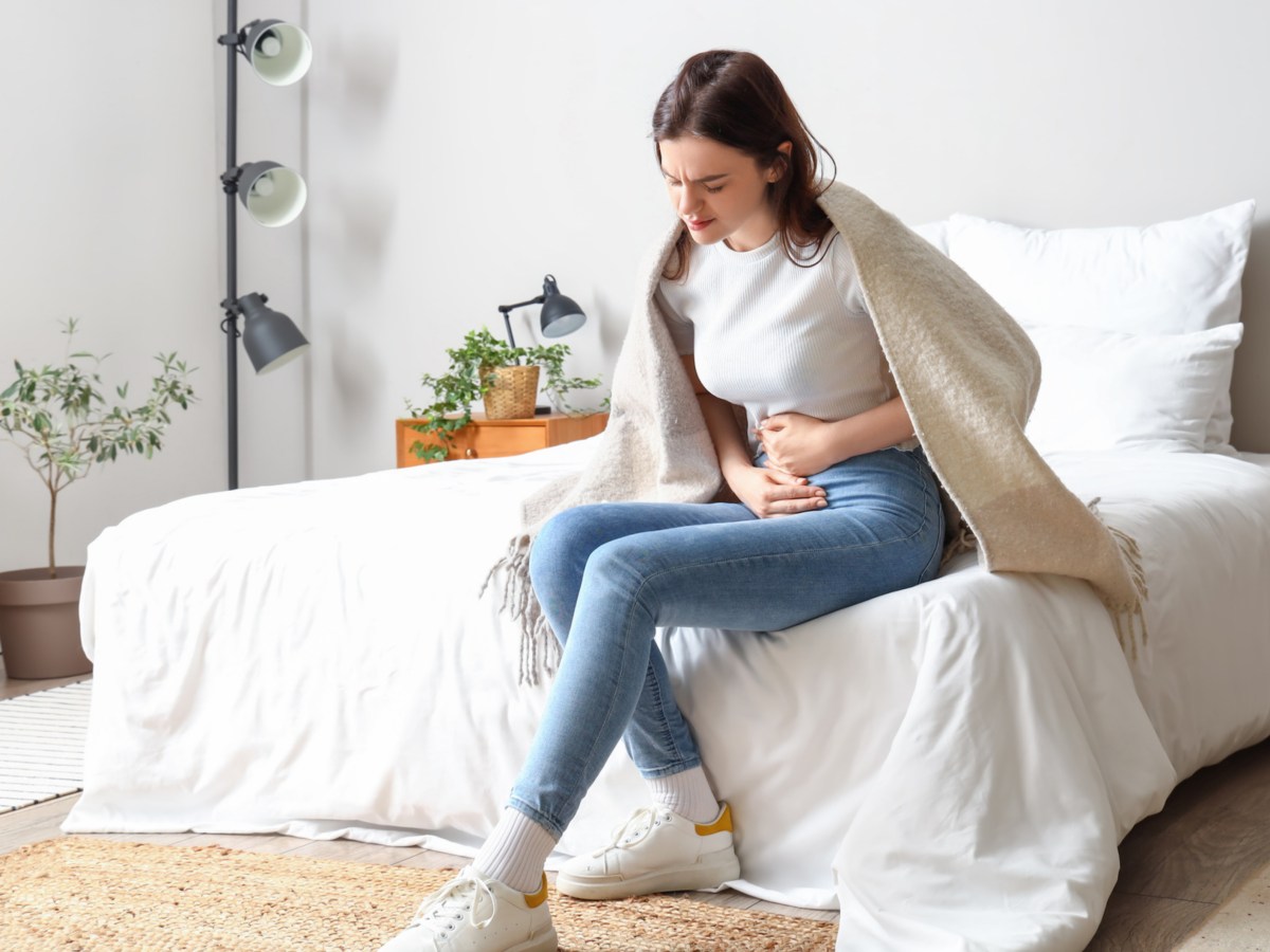 Frau auf einem Bett mit Decke über den Schultern, die sich mit schmerzendem Gesicht an den Bauch greift