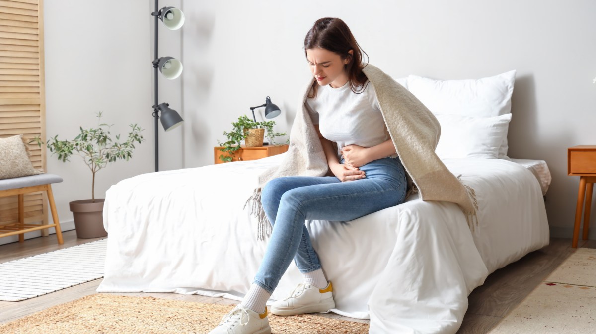Frau auf einem Bett mit Decke über den Schultern, die sich mit schmerzendem Gesicht an den Bauch greift