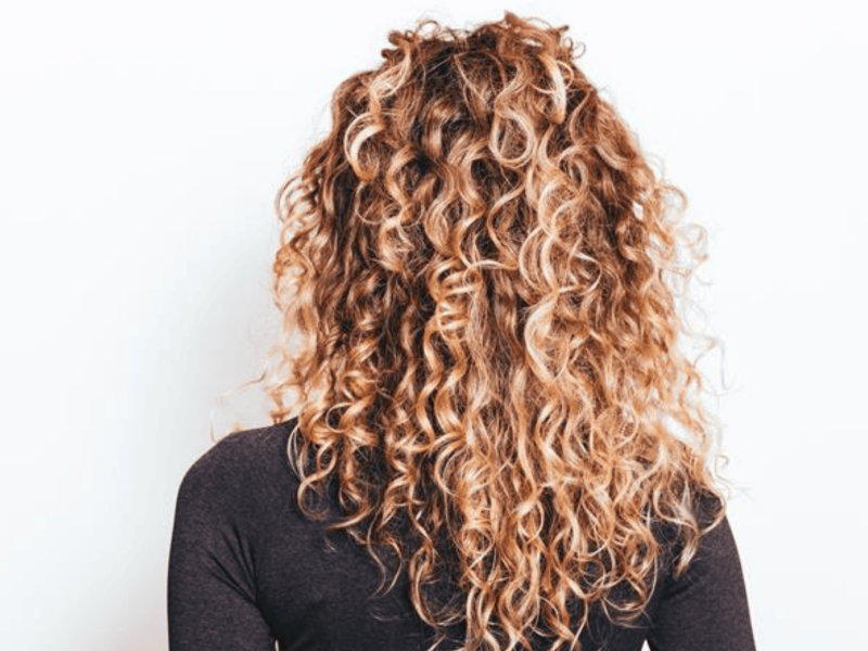 Junge frau mit langen, blonden Naturlocken steht mit dem r&uuml;cken zur kamera vor wei&szlig;er Wand in dunklem Shirt.