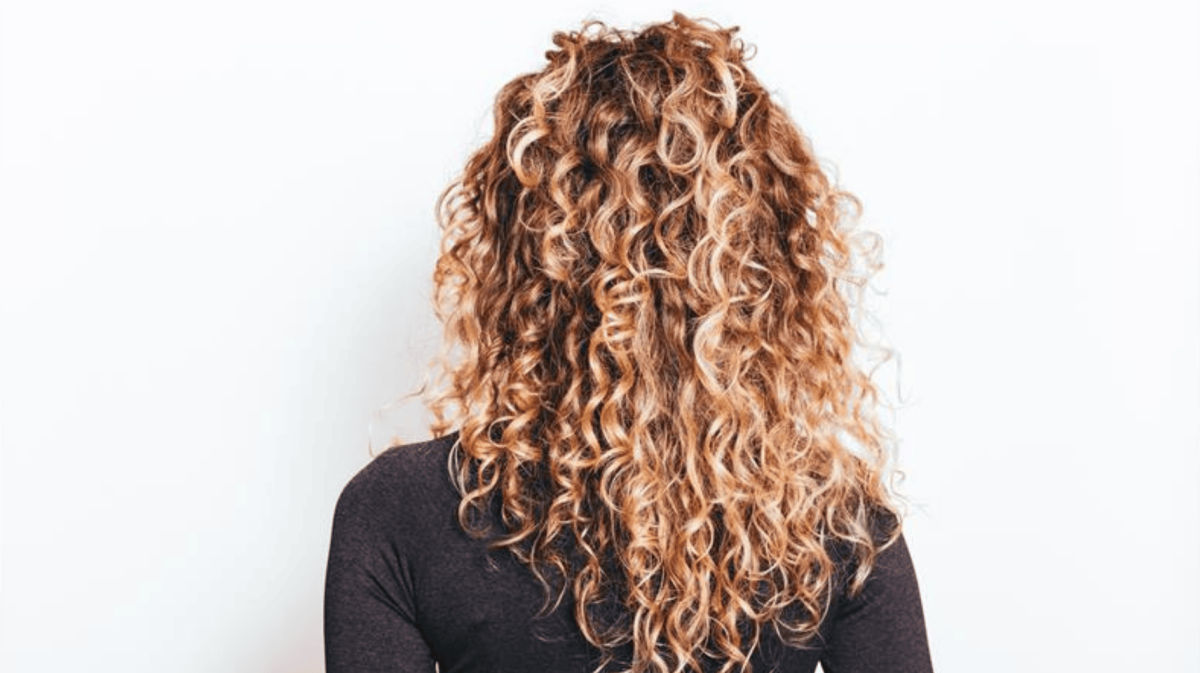 Junge frau mit langen, blonden Naturlocken steht mit dem rücken zur kamera vor weißer Wand in dunklem Shirt.