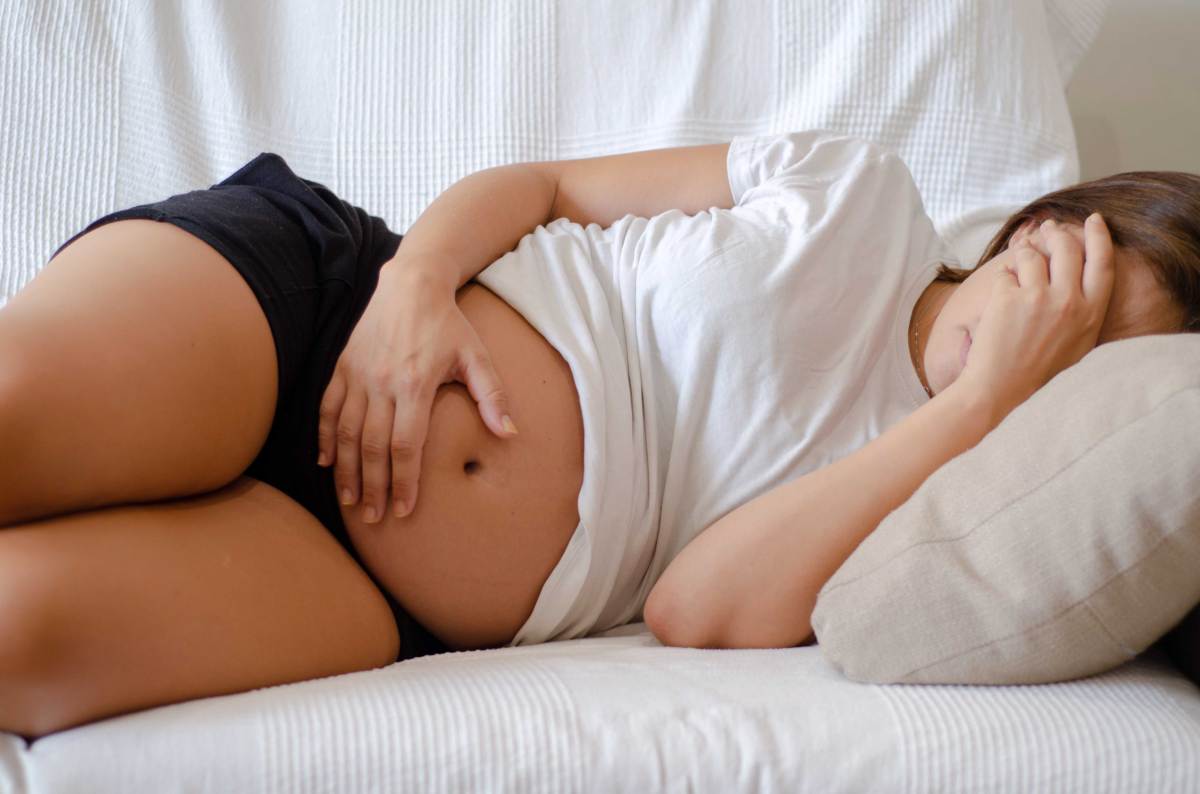 Schwangere Frau liegt auf dem Sofa, hält sich mit der einen Hand den Bauch, mit der anderen die Augen zu.