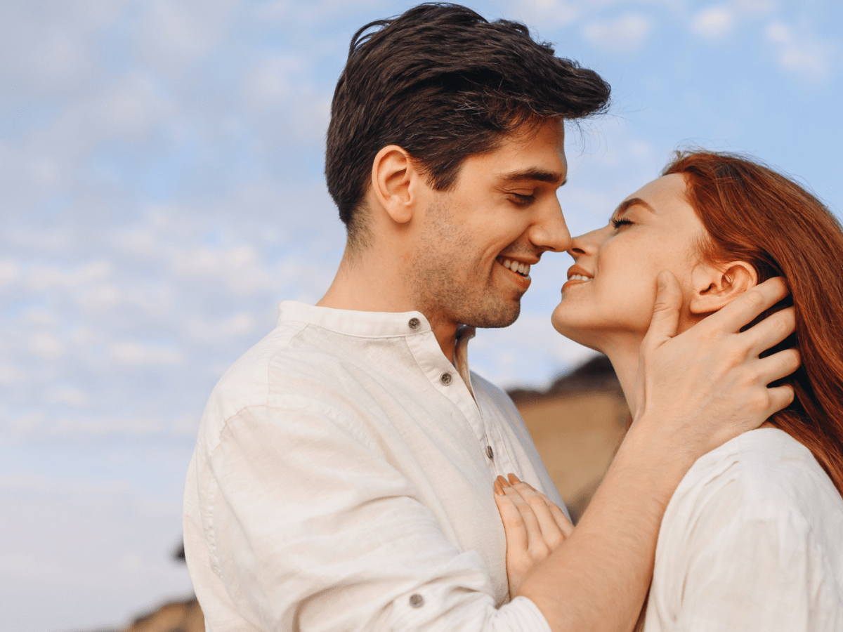 Ein Mann mit dunklem Haar und eine Frau mit roten langen Haaren stehen l&auml;chelnd nah beieinander unter klarem Himmel.