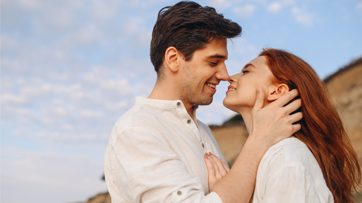 Ein Mann mit dunklem Haar und eine Frau mit roten langen Haaren stehen lächelnd nah beieinander unter klarem Himmel.