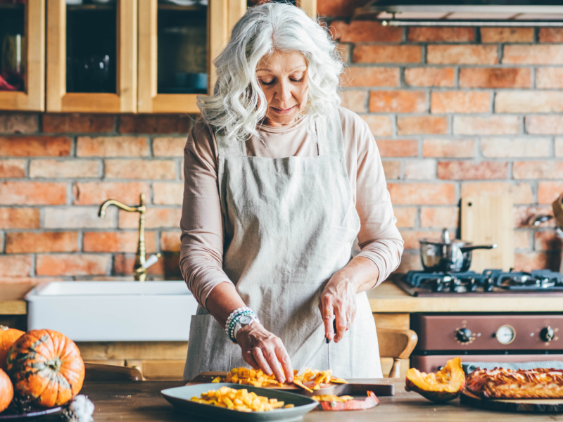 Ältere Frau in weißer Schürze schneidet gelben Kürbis gegen Küchengeräte zu Hause.