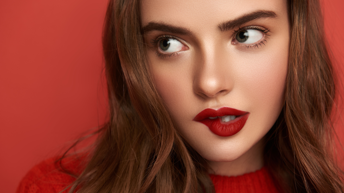 Junge Frau mit langen braunen Haaren, leuchtend roten Lippen und intensivem Blick vor einem roten Hintergrund.