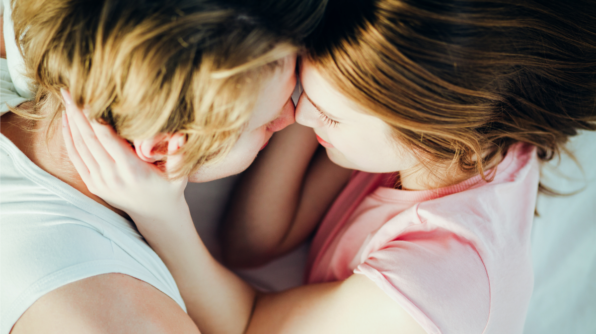 Junges Paar mit blonden Haaren liegt eng umschlungen, ihre Stirnen aneinander, sie lächeln zärtlich auf weißem Bett.