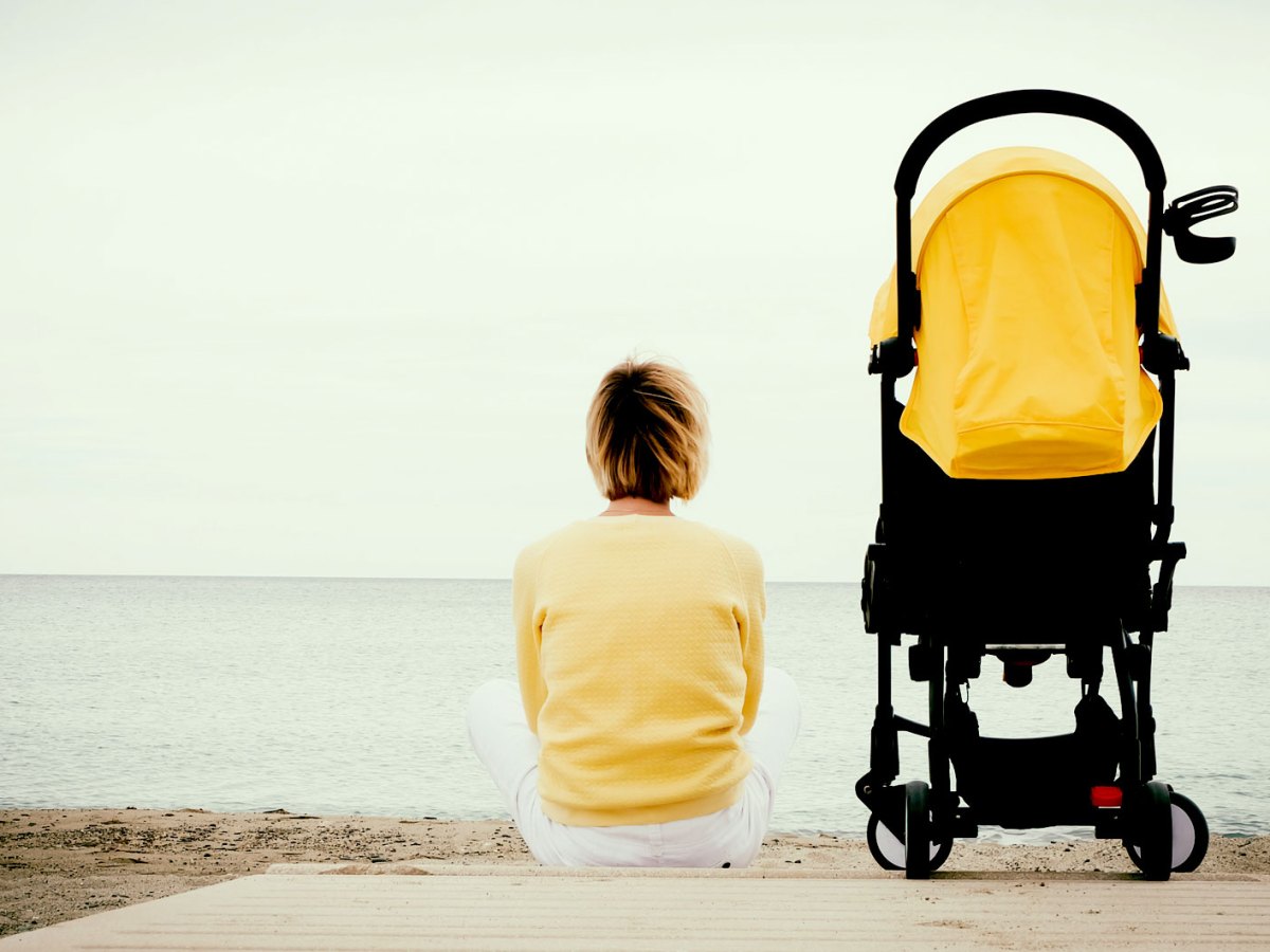 Frau im gelben Pullover sitzt auf einem Steg am Wasser, neben ihr ein gelber Kinderwagen.