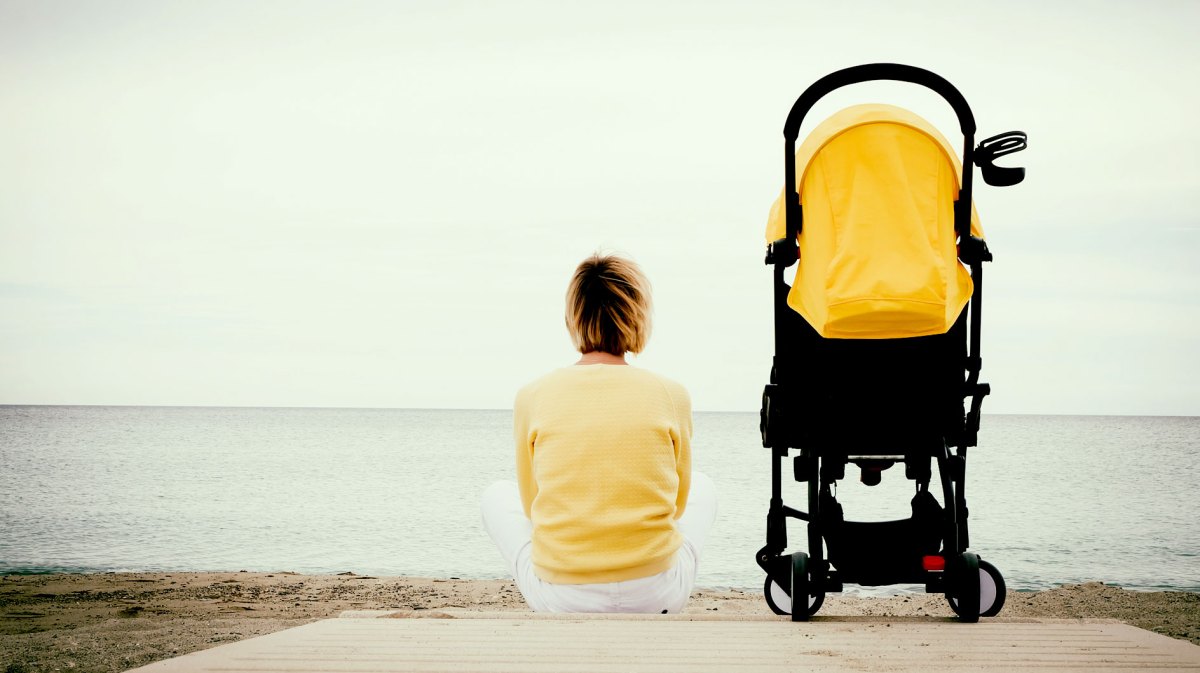 Frau im gelben Pullover sitzt auf einem Steg am Wasser, neben ihr ein gelber Kinderwagen.