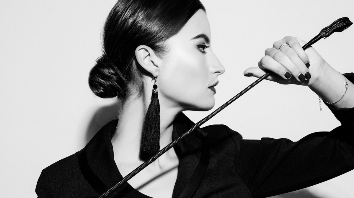 Elegante Frau mit strengem Dutt, langen schwarzen Ohrringen und Peitsche, blickt seitlich in dramatischer Schwarz-Weiß-Ästhetik. Junge Frau in schwarzem Blazer mit glänzenden Lippen und dunklen Nägeln hält selbstbewusst eine Peitsche vor weißem Hintergrund. Seitliches Porträt einer stilvollen Frau mit dezentem Make-up und Schmuck, die eine Peitsche in der Hand hält.