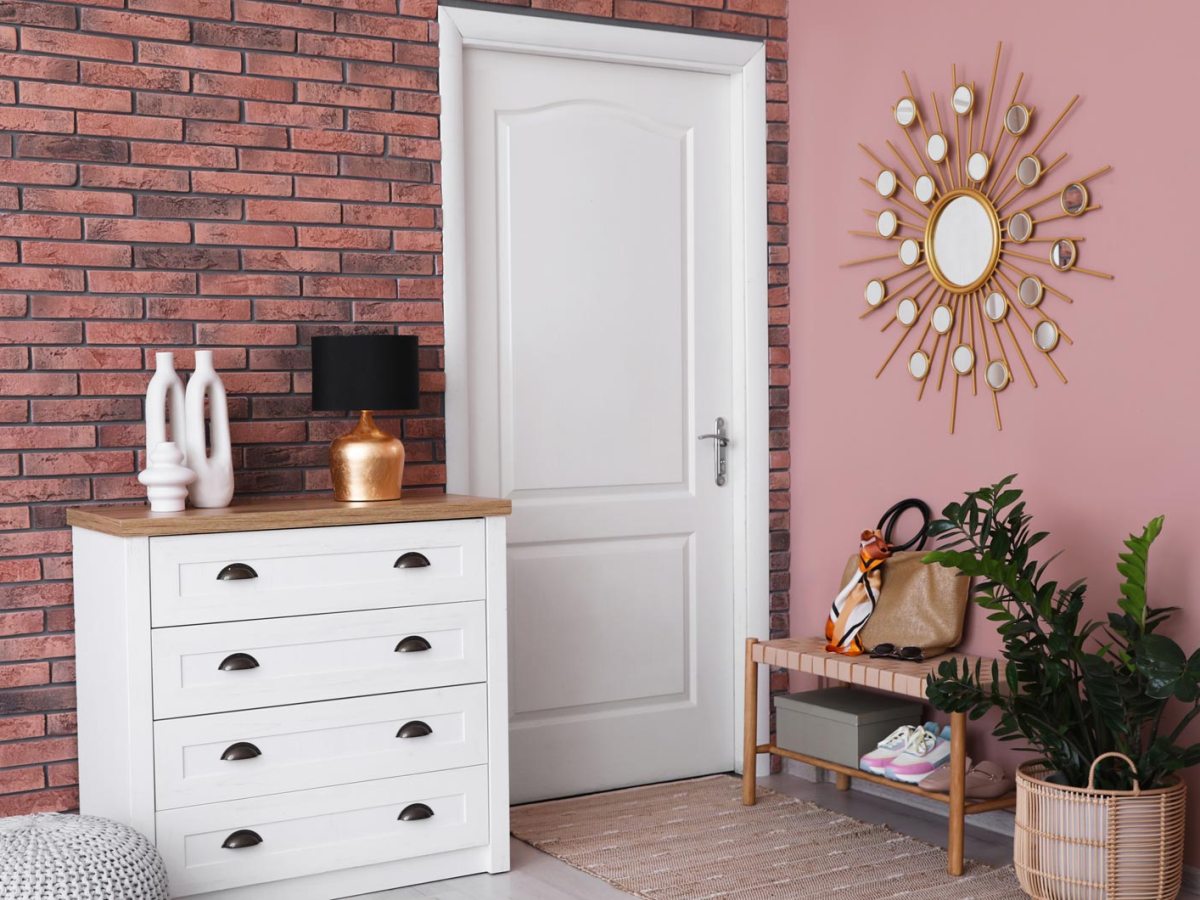 Heller Flur mit wei&szlig;er Kommode, goldener Lampe und Deko-Vasen vor roter Backsteinwand neben rosa Wand mit Sonnenspiegel.