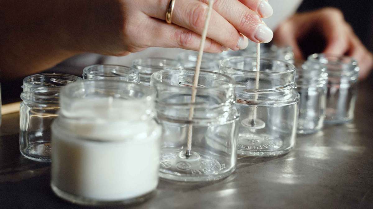 Eine Frau mit heller Haut setzt präzise Dochte in leere Gläser, umgeben von weißen Kerzen in einem sanft beleuchteten Raum.