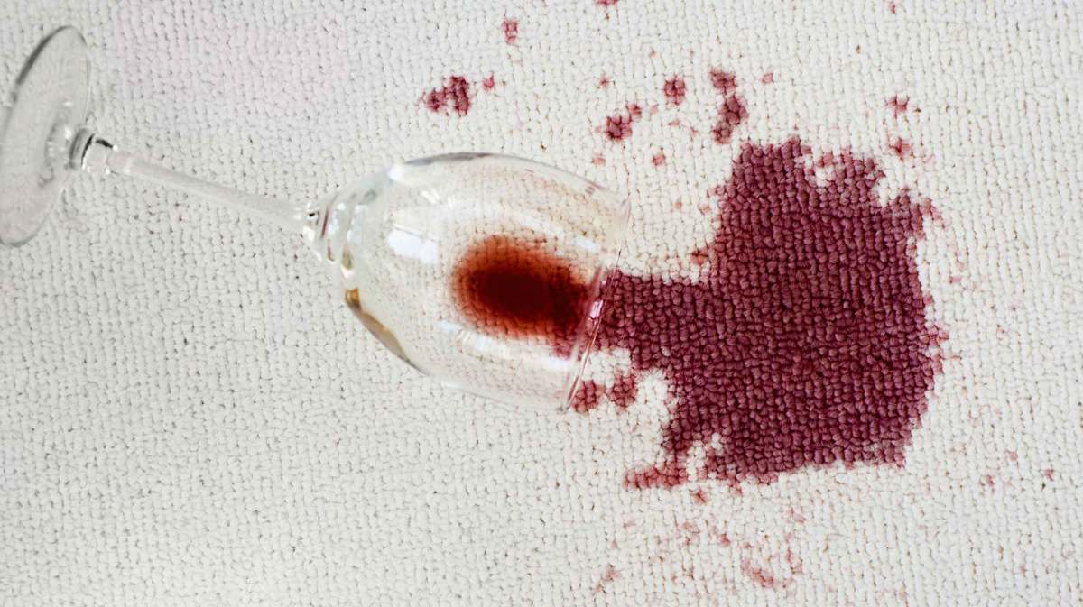Umgekipptes Rotweinglas liegt auf einem hellen Teppich mit einem großen Rotweinfleck.