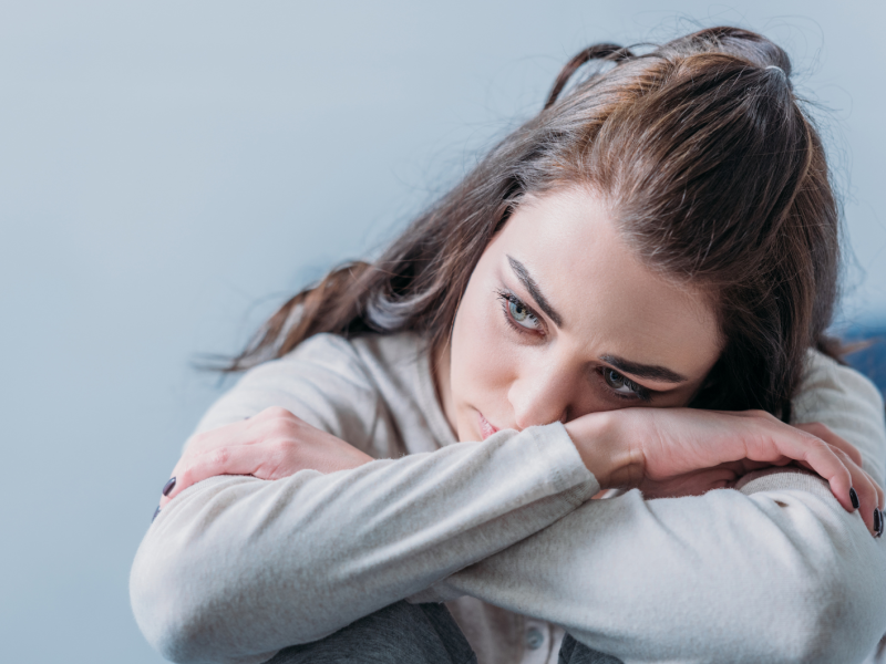 Dunkelhaarige junge Frau sitzt mit verschränkten Armen und traurigem Blick auf einem Stuhl.