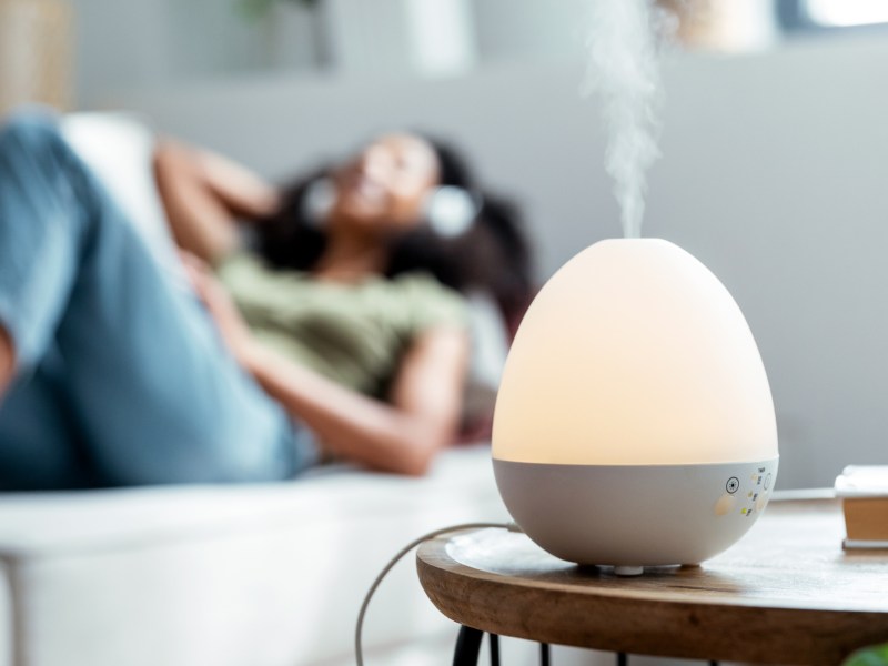 Eine Frau liegt auf dem Sofa. Vor ihr steht ein Raumluftbefeuchter, der die Luftfeuchtigkeit in der Wohnung erhöht.