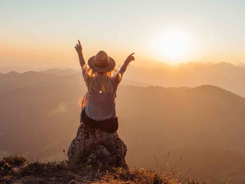Frau mit Hut sitzt auf einem Felsen vor einer Landschaft.