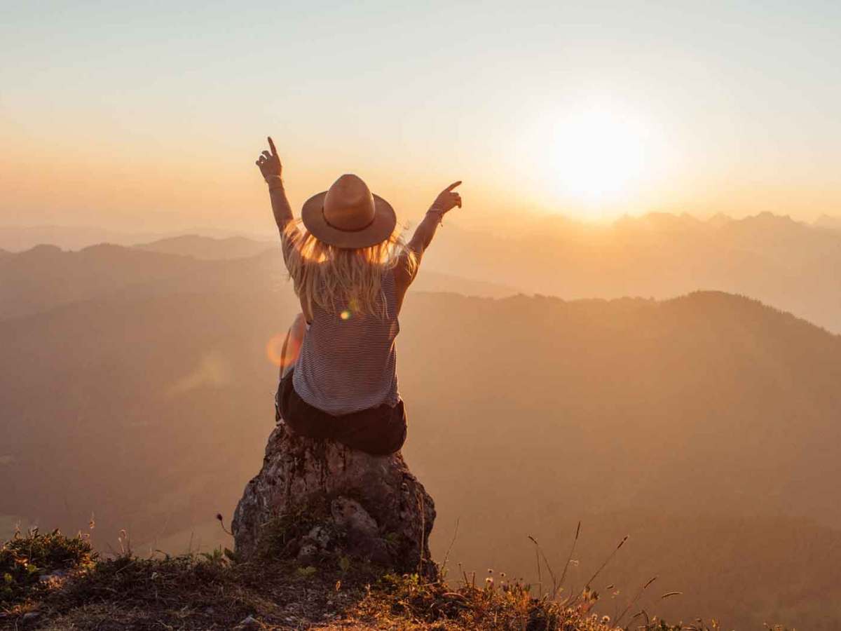 Frau mit Hut sitzt auf einem Felsen vor einer Landschaft.