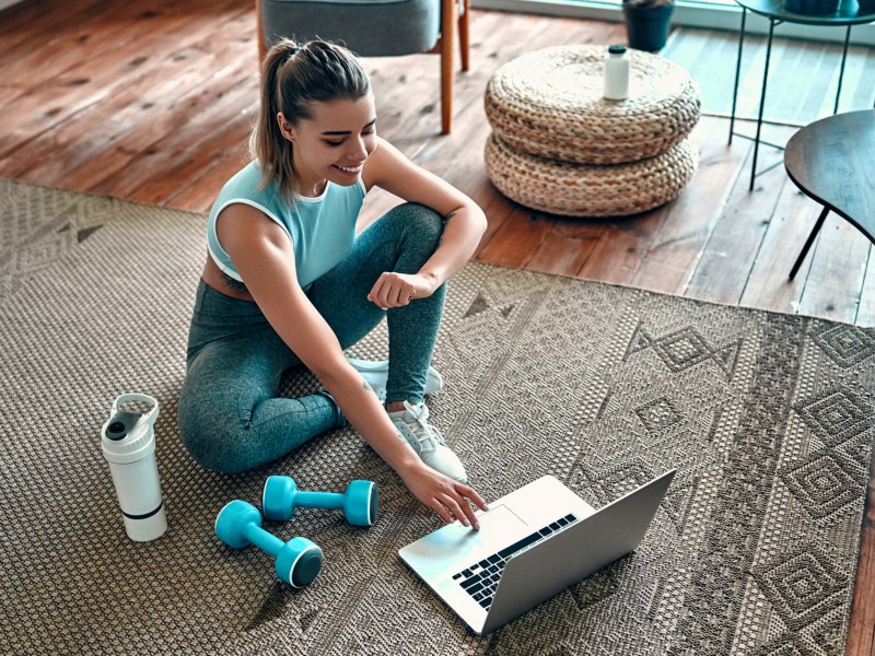 Frau sitzt mit Fitness-Geräten auf dem Boden vor ihrem Laptop