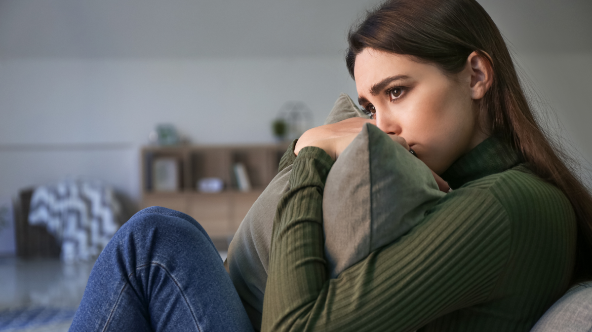 Dunkelhaarige junge Frau sitzt mit traurigem Blick und einem Kissen in der Hand auf dem Sofa