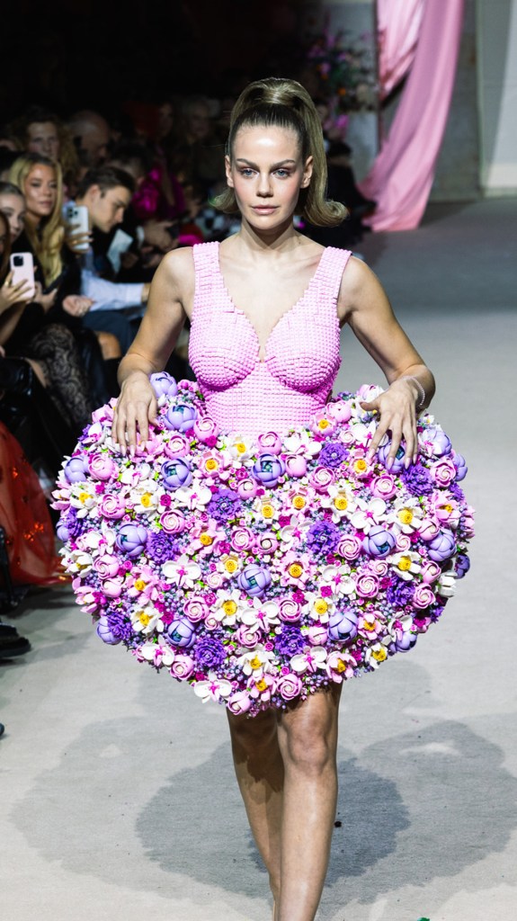 Ein Model mit strengem Pferdeschwanz trägt ein pinkes Kleid aus LEGO-Steinen mit herzförmigem Rock aus unzähligen bunten LEGO-Blüten auf dem Laufsteg.