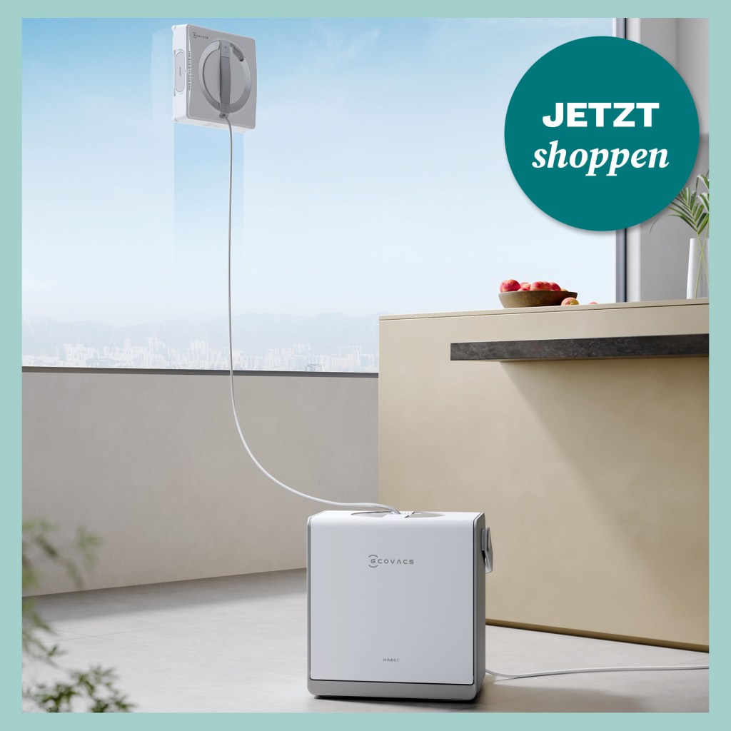 Moderner Fensterputzroboter saugt sich an klarer Glasfläche fest, daneben Küche mit Obstschale, dahinter Skyline im Dunst.