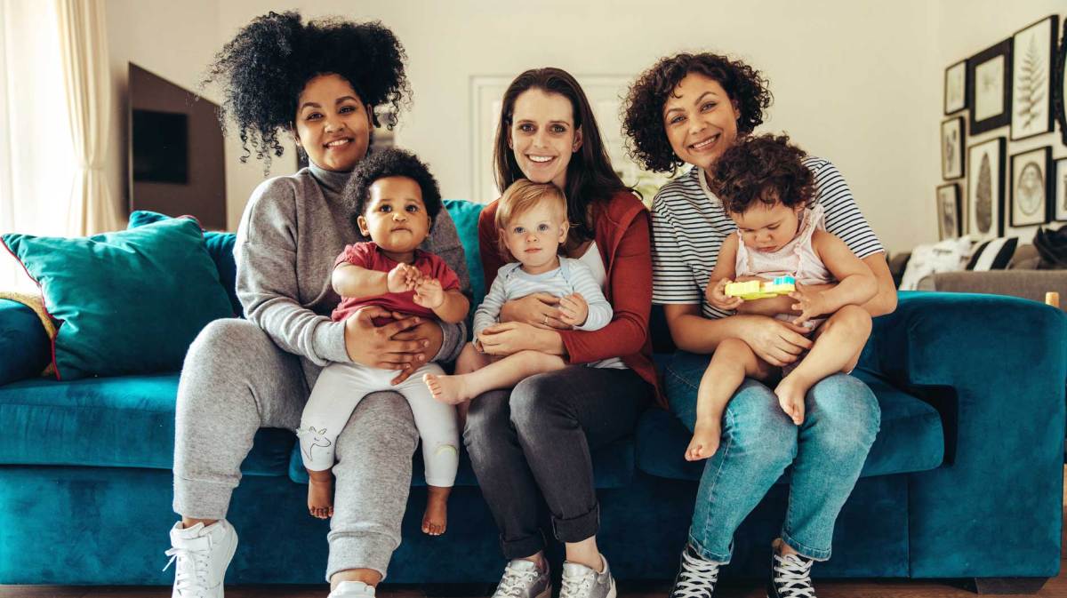 Drei Mamas haben ihre Kleinkinder auf dem Schoß, sitzen auf einem blauen Sofa und strahlen in die Kamera.