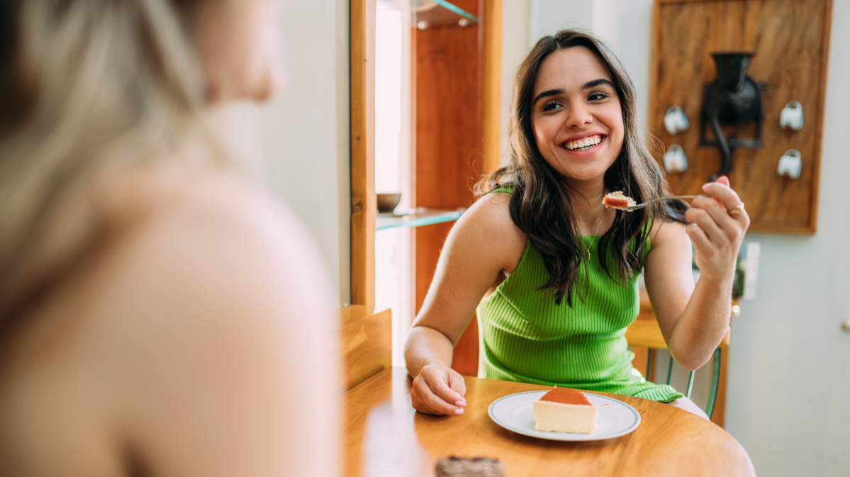 Eine junge Frau mit langen dunklen Haaren und grünem Top lächelt, während sie ein Stück Kuchen an einem Holztisch genießt.