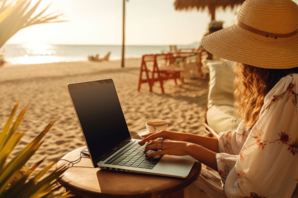 Frau sitzt am Strand mit dem Laptop auf dem Schoß.