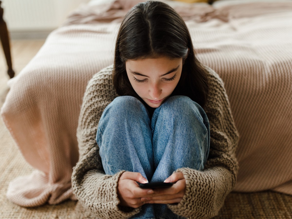 Frau, die vor ihrem Bett sitzt und schüchtern auf ihr Handy starrt