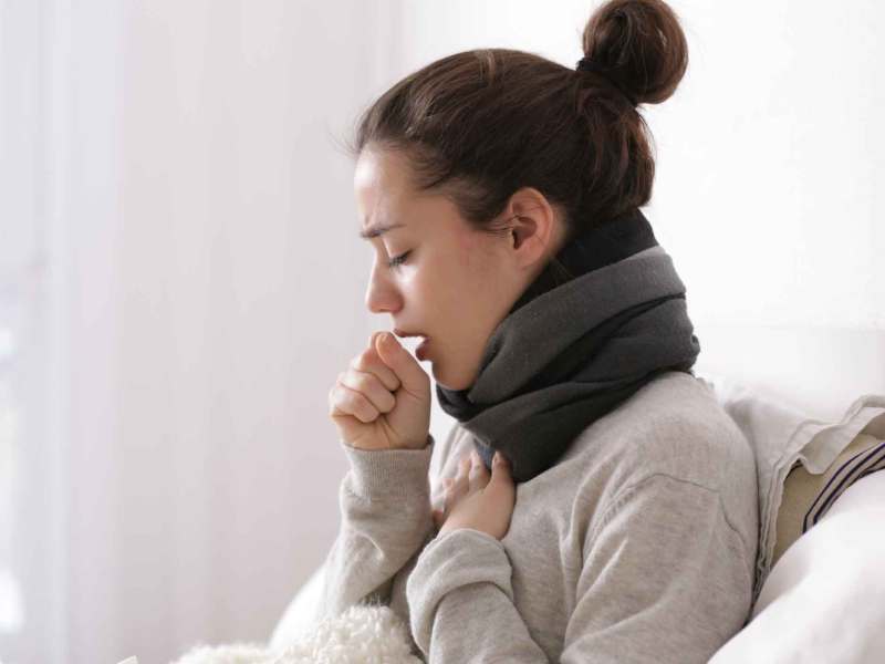 Frau mit Dutt sitzt auf dem Bett und hustet.