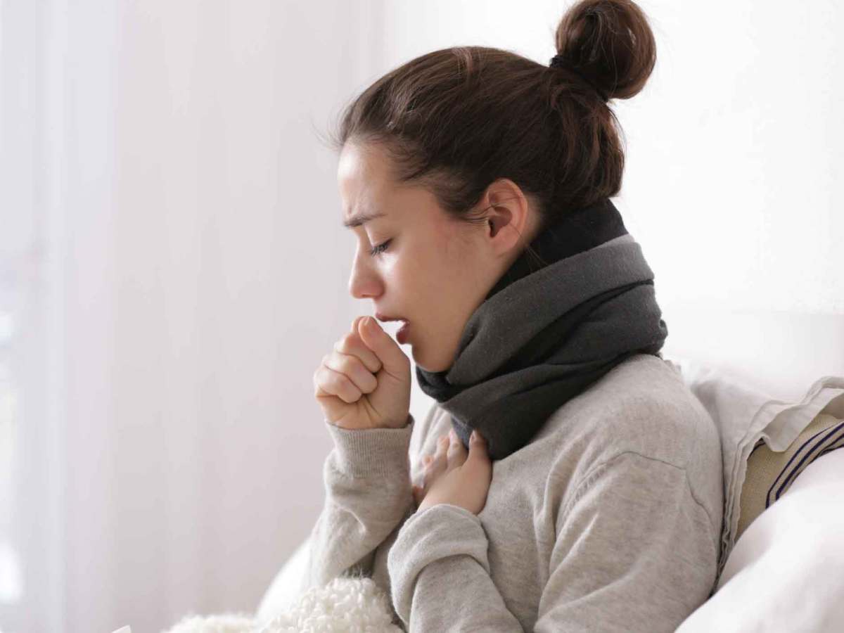 Frau mit Dutt sitzt auf dem Bett und hustet.