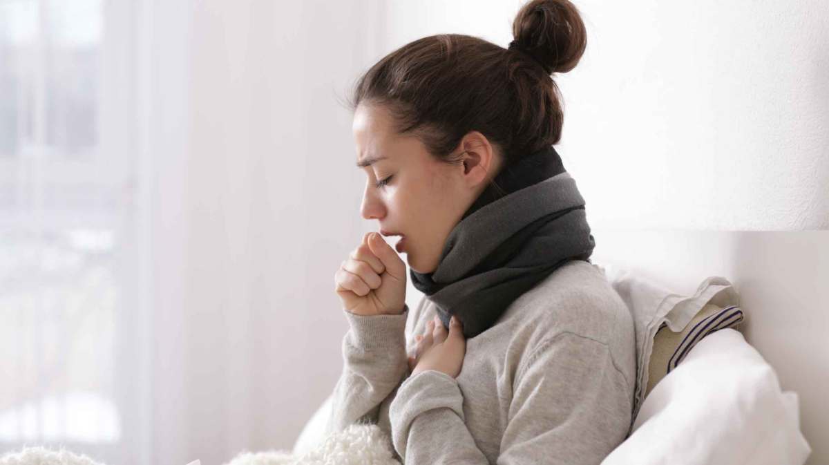 Frau mit Dutt sitzt auf dem Bett und hustet.