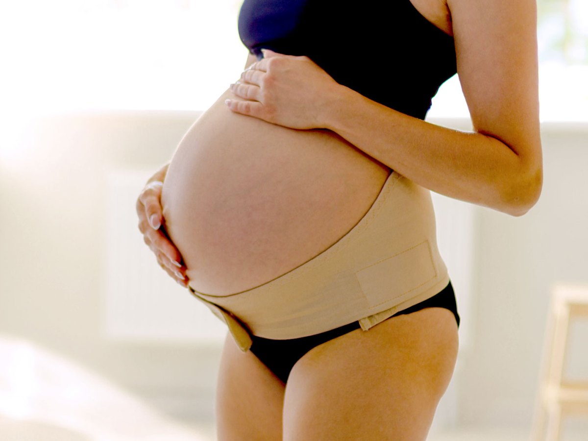 Hochschwangere Frau hat sich einen breiten Klettg&uuml;rtel um das Becken gelegt, welcher den gro&szlig;en Babybauch st&uuml;tzt.