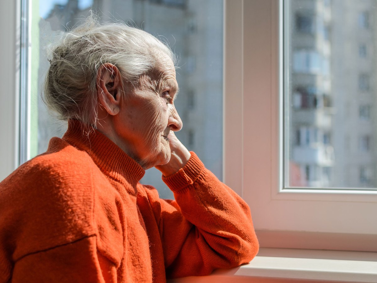 alte Frau sitzt in einem orangefarbenen Pullover am Fenster
