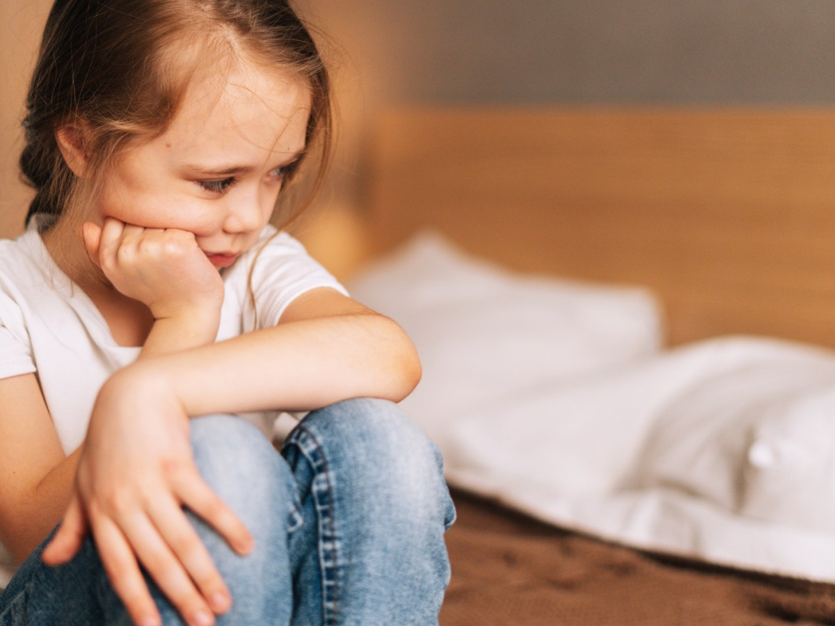 M&auml;dchen, ca. 6 Jahre, sitzt auf dem Bett und schaut besch&auml;mt zur Seite.