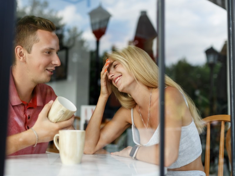Frau und Mann hinter Glasscheibe, die lachen und einen Kaffee aus einer Tasse trinken