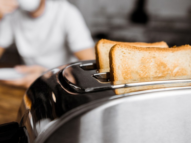 Ein Mann sitzt am Frühstückstisch. Im Vordergrund steht ein Toaster mit frisch getoasteten Brotscheiben.