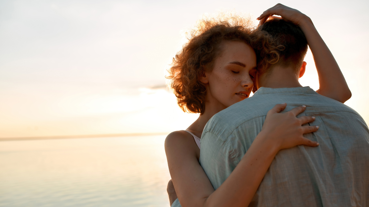 Pärchen am Wasser beim Sonnenuntergang, das sich liebevoll umarmt.