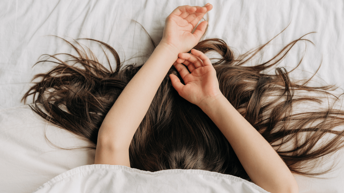 Frau im Bett, die sich unter der Decke versteckt und mit den Armen und dunkelbraunen Haaren herausguckt.