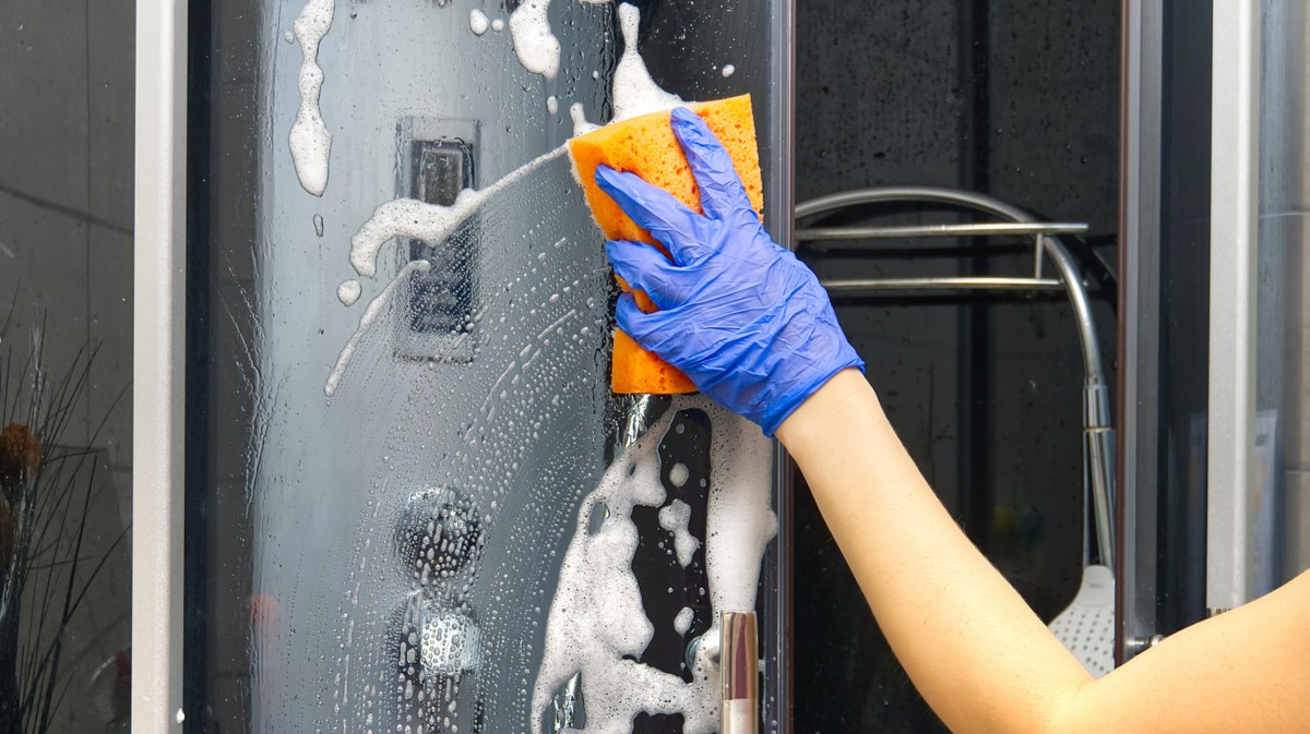 Person putzt mit einem blauen Gummihandschuh und einem orangen Schwamm die Duschkabine.