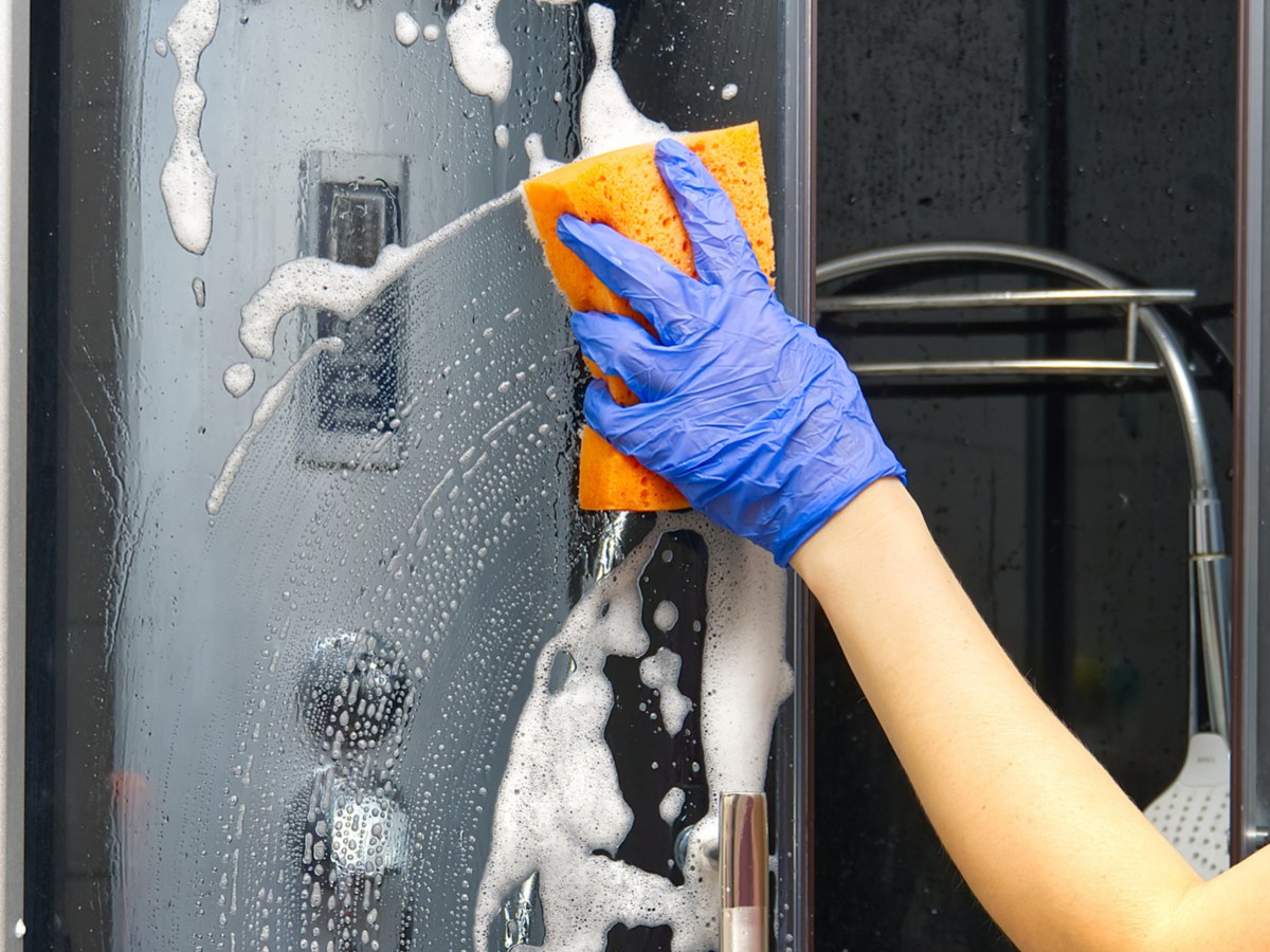 Person putzt mit einem blauen Gummihandschuh und einem orangen Schwamm die Duschkabine.