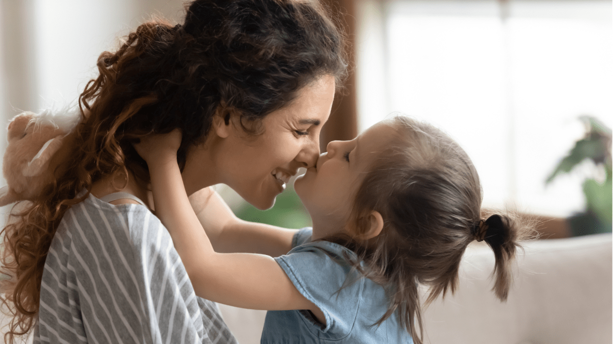 Mutter hält ihre Tochter vor sich, die sie auf die Nase küsst.