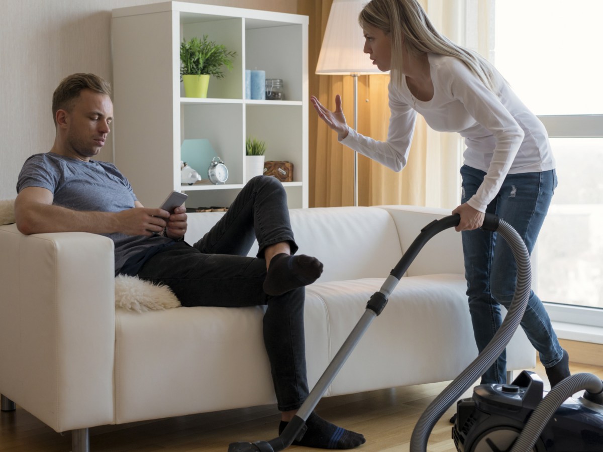 Mann sitzt auf Sofa während Frau staubsaugt.