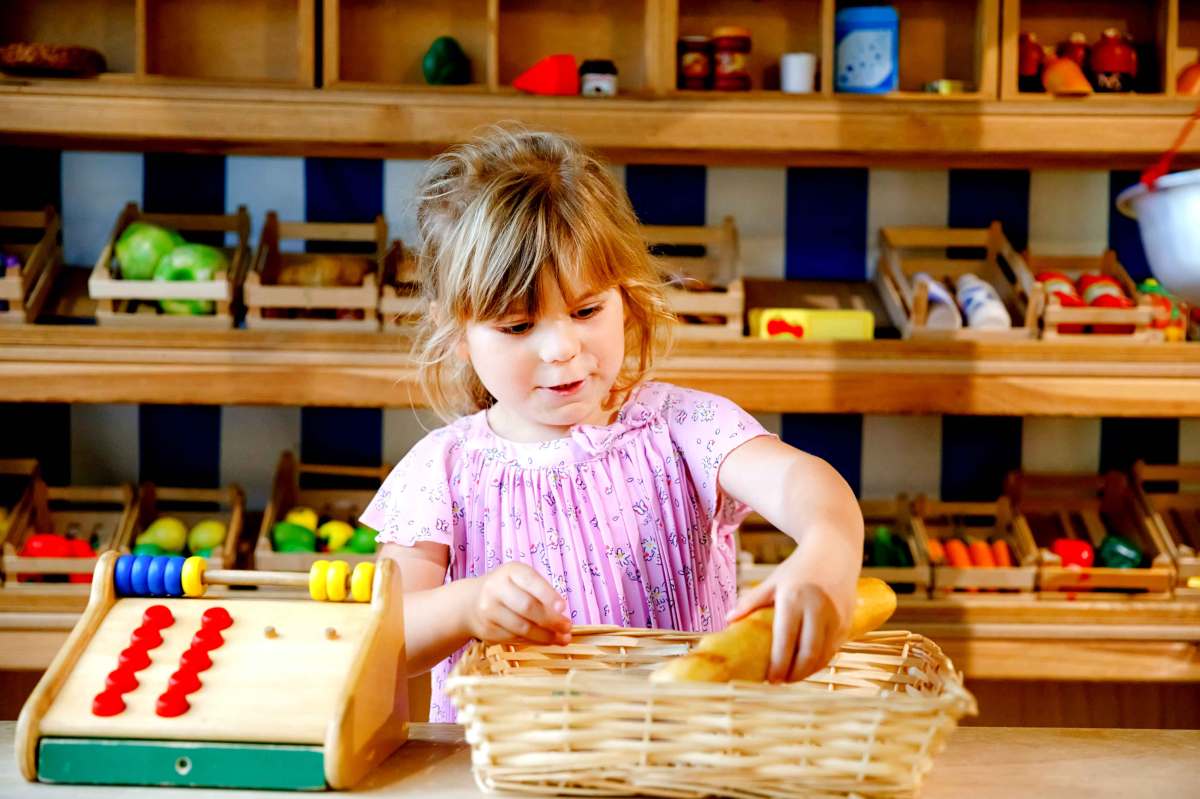 Kleines Mädchen steht in ihrer Holzspielküche und legt ein Spielzeugbaguette in einen Korb.