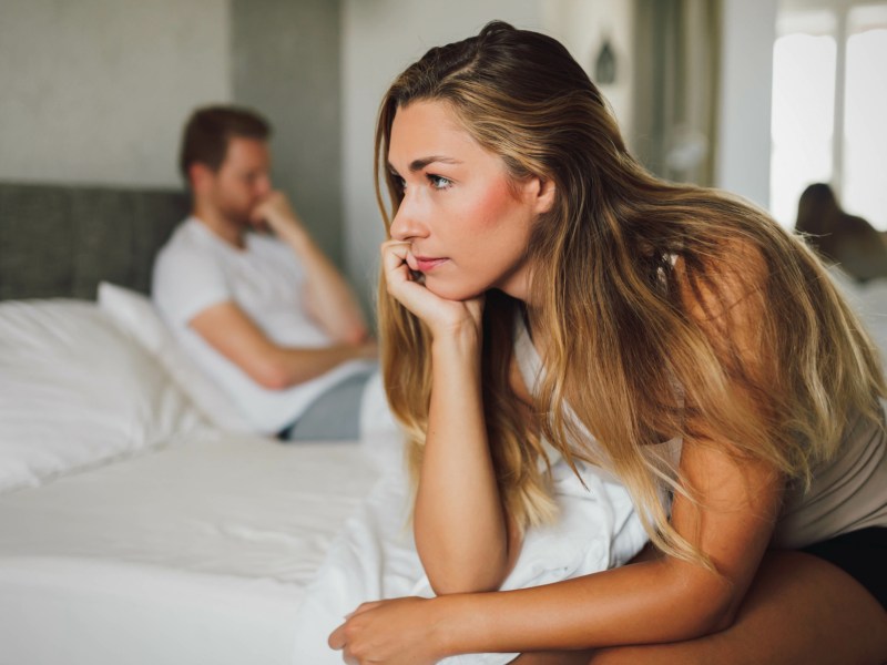 Frau sitzt im Vordergrund auf der Bettkante und starrt ins Leere, während ein Mann im Hintergrund auf dem Bett sitzt und nachdenklich nach vorne schaut