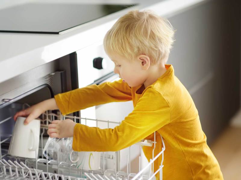 Junge um die vier Jahre räumt die Spülmaschine ein.
