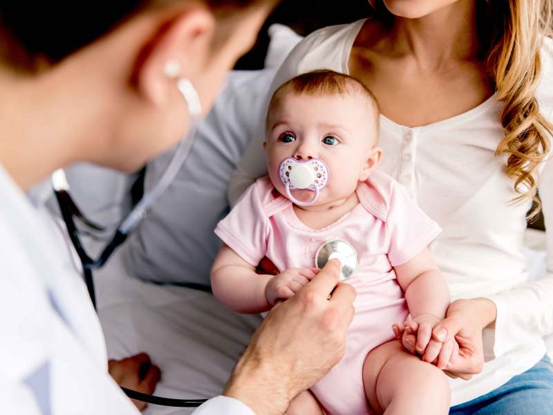 Baby sitzt auf dem Schoß der Mutter und schaut den Arzt mit großen Augen an, der es mit einem Stethoskop abhören möchte,